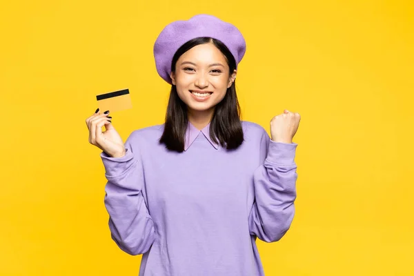 Excitada mujer asiática en boina celebración de tarjeta de crédito y regocijo aislado en amarillo - foto de stock