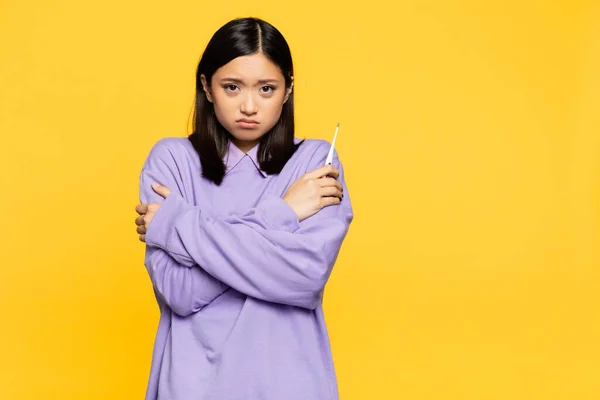 Sick asian woman holding digital thermometer while freezing isolated on yellow — Stock Photo