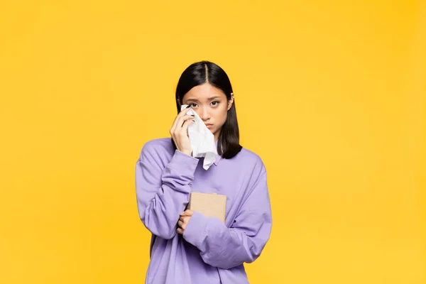 Sad asian woman holding tissue box and napkin while crying isolated on yellow — Stock Photo