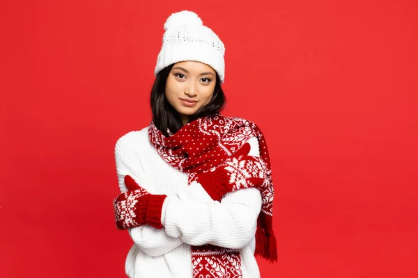 Cheerful asian woman in mittens, hat and scarf embracing herself isolated on red — Stock Photo