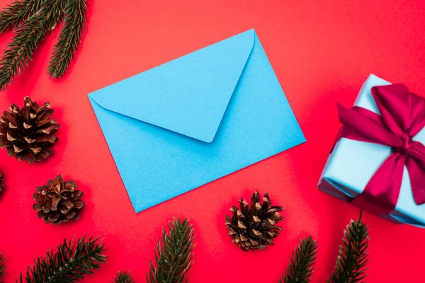 Vista dall'alto della confezione regalo, coni, rami di abete rosso e busta blu su sfondo rosso — Foto stock