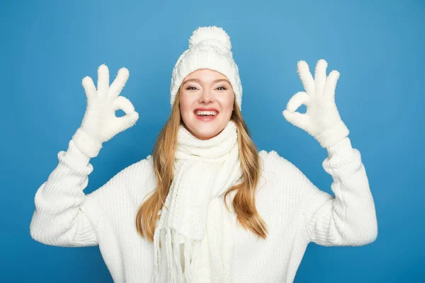 Счастливая блондинка красивая женщина в зимнем белом наряде, показывающем ОК на синем фоне — стоковое фото