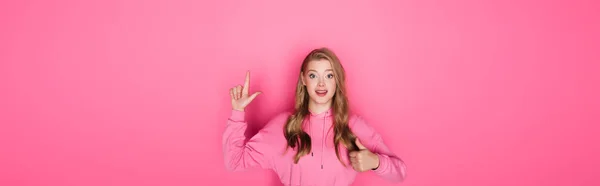 Sorrindo bela mulher apontando com o dedo e mostrando o polegar no fundo rosa, banner — Fotografia de Stock