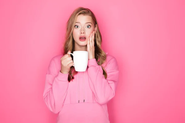 Schockierte schöne Frau mit offenem Mund und Becher auf rosa Hintergrund — Stockfoto
