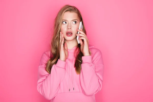 Потрясенная красивая женщина с открытым ртом разговаривает на смартфоне на розовом фоне — стоковое фото