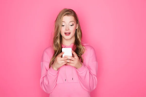 Збуджена красива жінка з відкритим ротом тримає смартфон на рожевому фоні — стокове фото