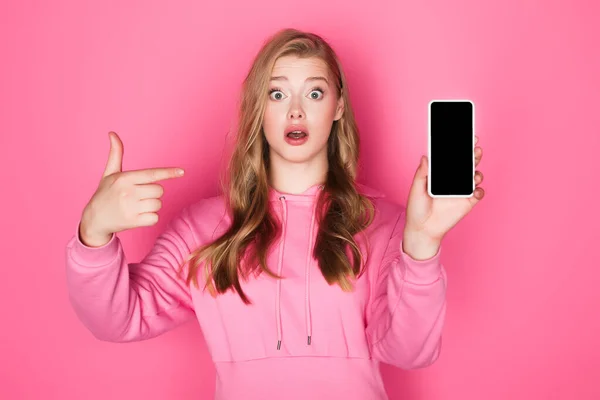 Shocked beautiful woman pointing at smartphone on pink background — Stock Photo