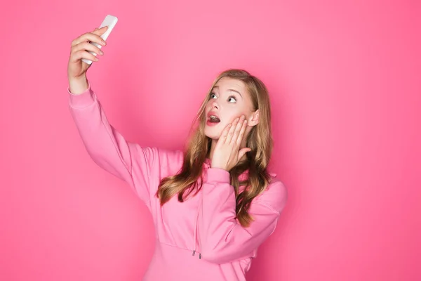 Шокована красива жінка бере селфі на смартфон на рожевому фоні — стокове фото