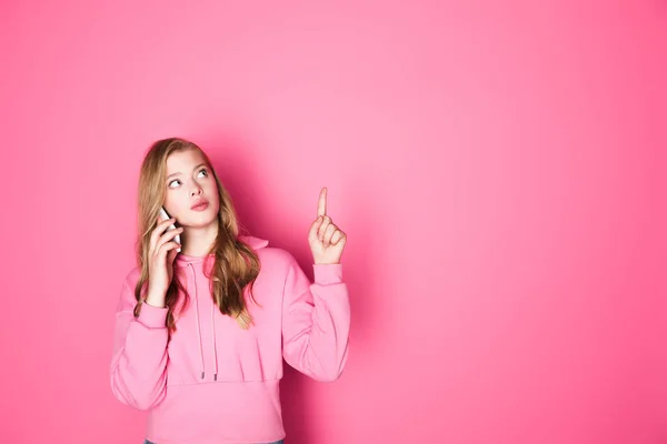 Hermosa mujer hablando en el teléfono inteligente y apuntando hacia arriba en el fondo rosa - foto de stock