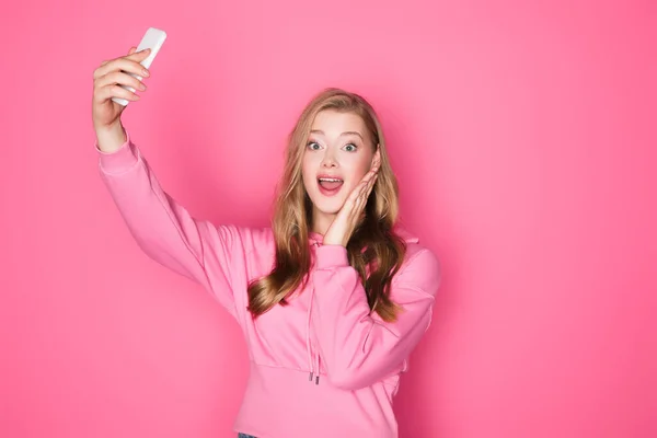 Shocked beautiful woman taking selfie on smartphone on pink background — Stock Photo