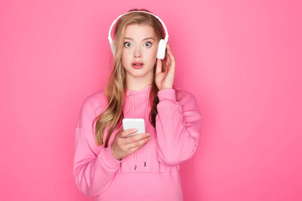 Shocked beautiful woman in headphones with smartphone on pink background — Stock Photo