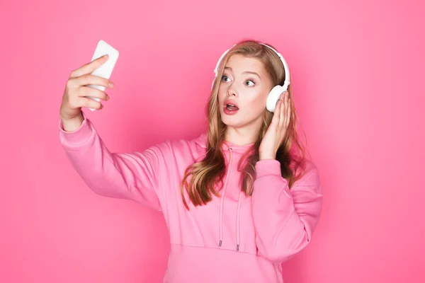 Shocked beautiful woman in headphones taking selfie on pink background — Stock Photo