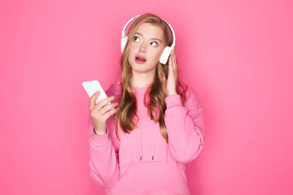 Hermosa mujer pensativa en auriculares con teléfono inteligente sobre fondo rosa - foto de stock