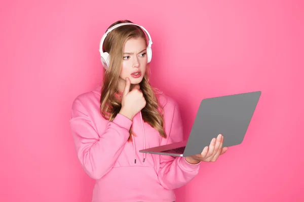 Hermosa mujer pensativa en auriculares con portátil sobre fondo rosa - foto de stock