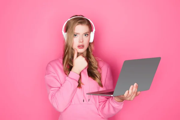 Mulher bonita pensativa em fones de ouvido com laptop no fundo rosa — Fotografia de Stock