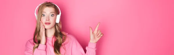 Impactado hermosa mujer escuchando música en los auriculares y apuntando hacia arriba en el fondo rosa, pancarta - foto de stock
