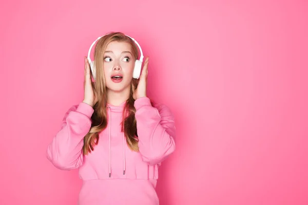 Impactado hermosa mujer escuchando música en auriculares sobre fondo rosa - foto de stock