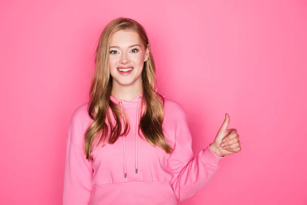 Heureux belle jeune femme en sweat à capuche montrant pouce vers le haut sur fond rose — Photo de stock