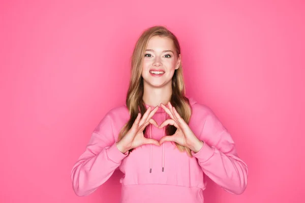 Schöne junge Frau im Kapuzenpullover zeigt Herz mit Händen auf rosa Hintergrund — Stockfoto