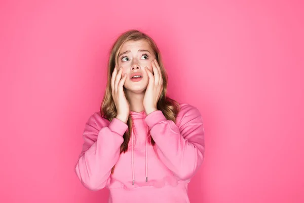 Shocked beautiful young woman in hoodie touching face on pink background — Stock Photo