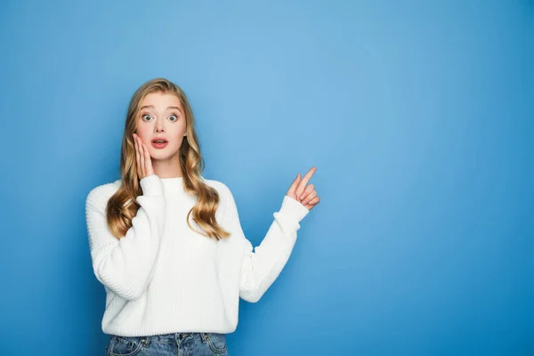 Scioccato bella donna bionda in maglione che punta con il dito isolato su blu — Foto stock