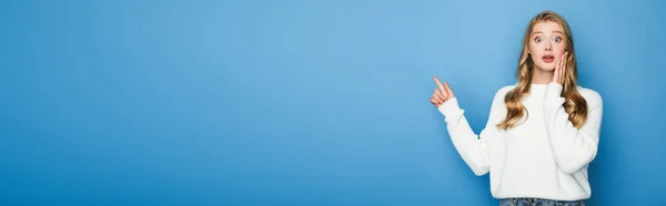Шокована блондинка красива жінка в светрі, вказуючи пальцем ізольована на синьому, банер — стокове фото