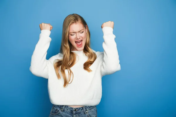 Happy blonde beautiful woman in sweater showing yeah gesture isolated on blue background — Stock Photo