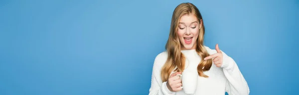 Happy blonde beautiful woman in sweater pointing at mug isolated on blue background, banner — Stock Photo