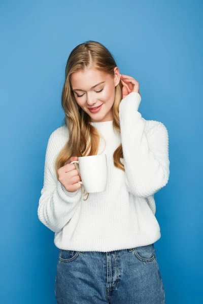 Happy blonde beautiful woman in sweater pointing at mug isolated on blue background — Stock Photo