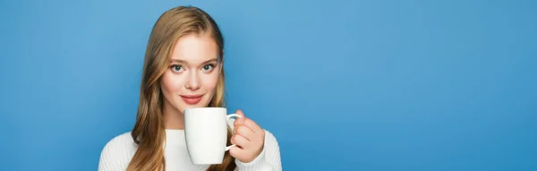 Smiling blonde beautiful woman in sweater with mug isolated on blue background, banner — Stock Photo