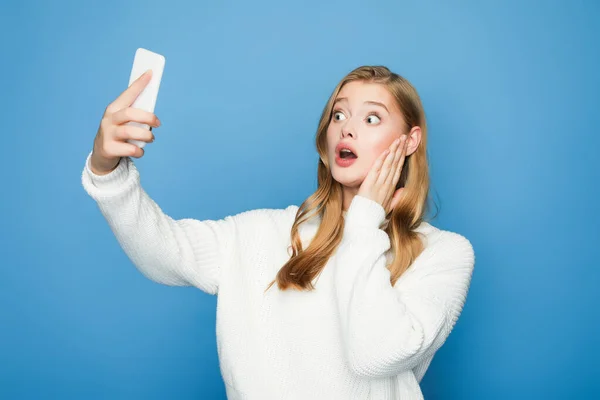 Impactado rubia hermosa mujer tomando selfie aislado en azul fondo - foto de stock