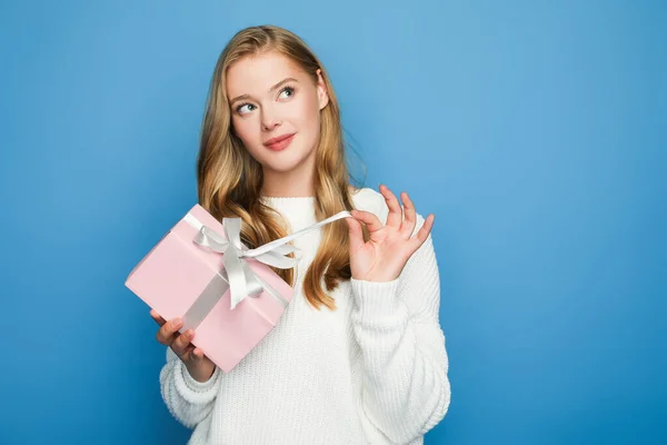 Mujer hermosa rubia soñadora en suéter con caja de regalo aislada en azul - foto de stock