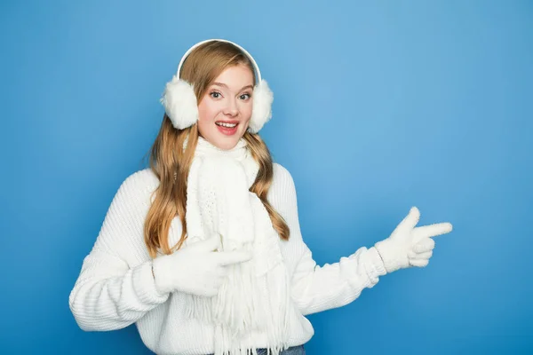 Усміхнена красива жінка в зимовому білому вбранні, вказуючи в сторону ізольована на синьому — стокове фото