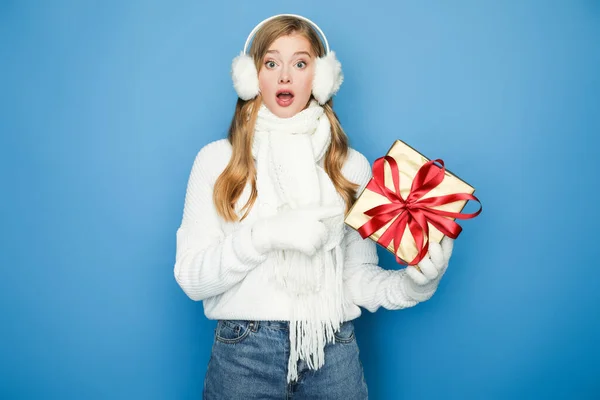 Здивована красива жінка в зимовому білому вбранні з подарунковою коробкою ізольована на синьому — стокове фото