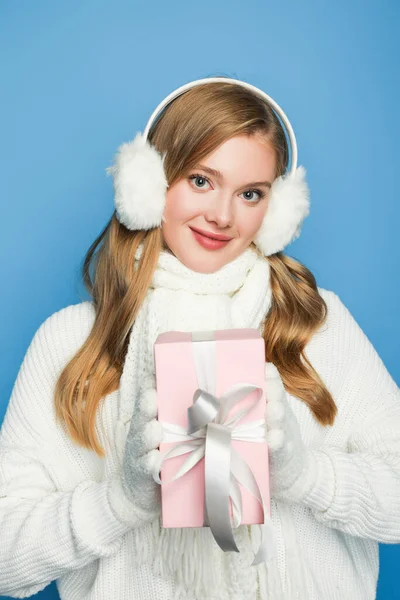 Улыбающаяся красивая женщина в зимнем белом наряде с подарочной коробкой изолированы на синий — стоковое фото