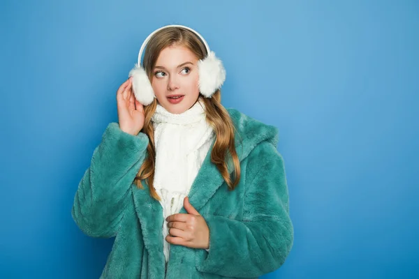 Stylish woman in green faux fur coat and ear muffs isolated on blue — Stock Photo