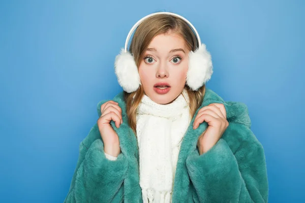 Surprised stylish woman in green faux fur coat and ear muffs isolated on blue — Stock Photo