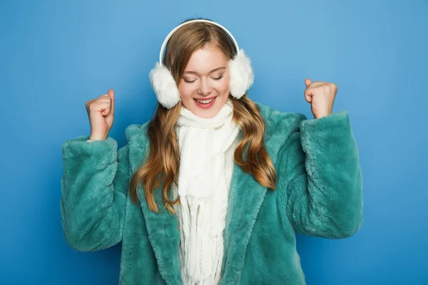 Glücklich stylische Frau in grünem Kunstpelzmantel und Ohrenschützer isoliert auf blau — Stockfoto