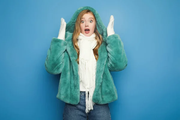 Shocked stylish woman in green faux fur coat looking away isolated on blue — стоковое фото