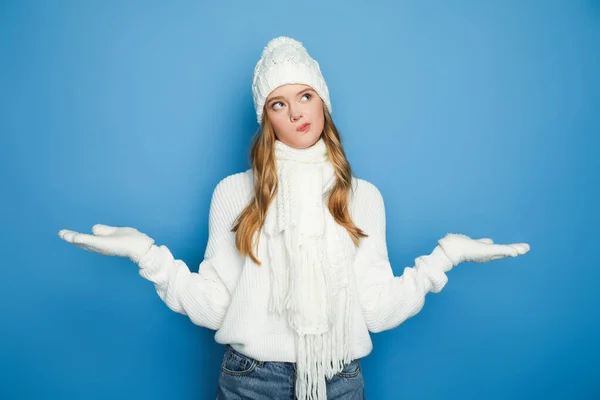 Verwirrte schöne Frau im winterweißen Outfit zeigt Achselzucken vereinzelt auf blauem Grund — Stockfoto