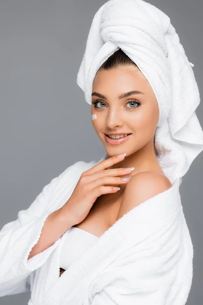 Усміхнена жінка з рушником на голові та косметичним кремом на обличчі ізольована на сірому — стокове фото