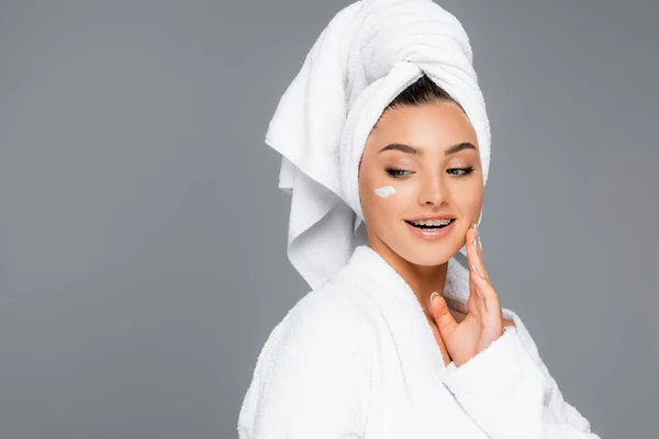 Donna sorridente con asciugamano sulla testa e crema cosmetica sul viso isolata sul grigio — Foto stock