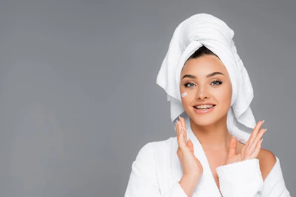 Femme souriante avec serviette sur la tête et crème cosmétique sur le visage isolé sur gris — Photo de stock