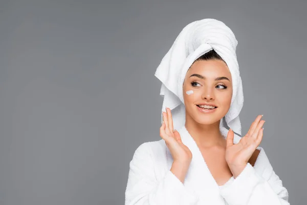 Mujer sonriente con toalla en la cabeza y crema cosmética en la cara aislada en gris - foto de stock