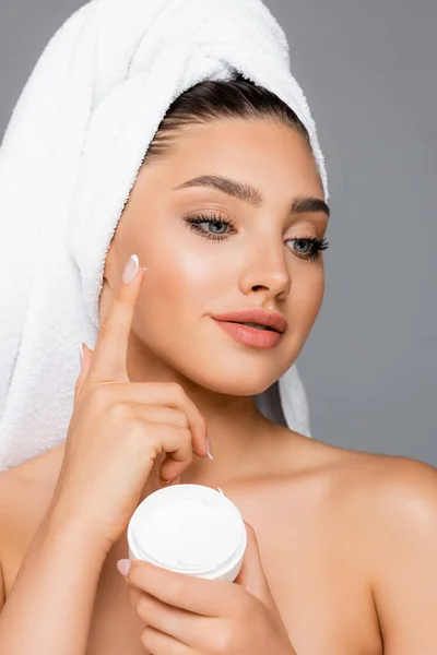 Donna con asciugamano sulla testa applicando crema cosmetica sul viso isolato su grigio — Foto stock