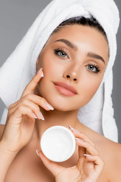 Donna con asciugamano sulla testa applicando crema cosmetica sul viso isolato su grigio — Foto stock