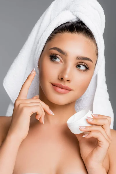 Donna con asciugamano sulla testa applicando crema cosmetica sul viso isolato su grigio — Foto stock