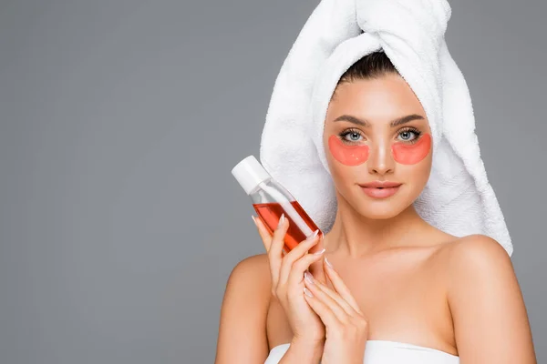 Woman with towel on head and eye patches holding lotion isolated on grey — Stock Photo