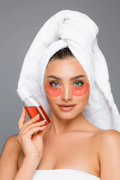 Woman with towel on head and eye patches holding lotion isolated on grey — Stock Photo