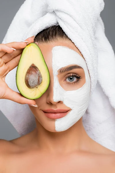 Donna con asciugamano sulla testa e maschera di argilla sul viso tenuta avocado isolato su grigio — Foto stock